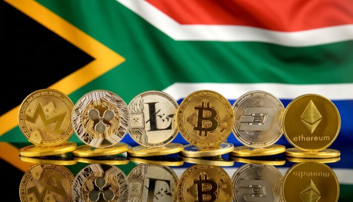 Physical cryptocurrency coins displayed against a reflective surface in front of the South African national flag