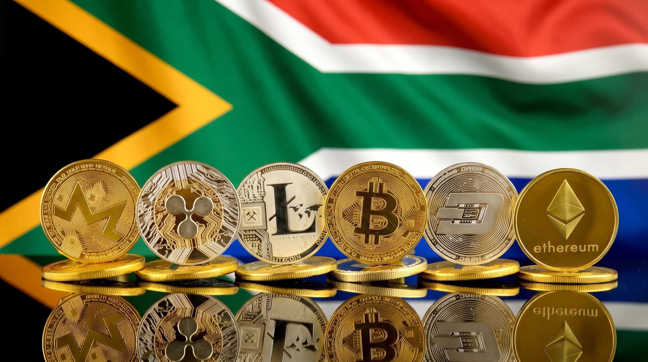 Physical cryptocurrency coins displayed against a reflective surface in front of the South African national flag